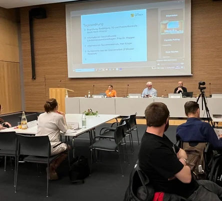 Perspektive auf Podium seitlich aus dem Podium / Tische mit Personen vor dem Podium , im Podium sitzen links ein Frau und in der Mitte und rechts zwei Männer