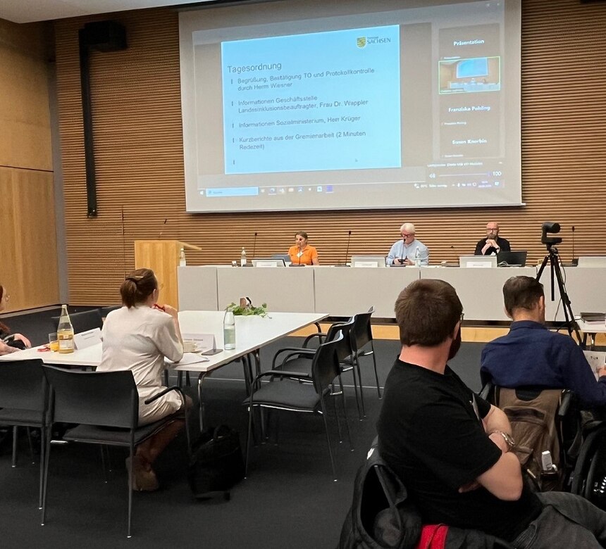 Perspektive auf Podium seitlich aus dem Podium / Tische mit Personen vor dem Podium , im Podium sitzen links ein Frau und in der Mitte und rechts zwei Männer