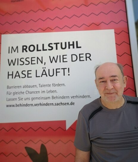 älterer Mann mit wenig Haaren steht vor dem Plakat behindern verhindern