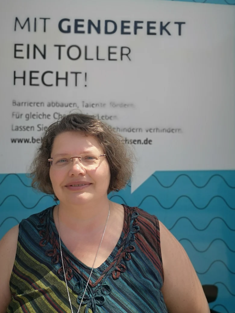 Frau mit halblangen gewellten Haaren und Brille ist mit Oberkörper zu sehen vor dem Plakat behindern verhindern