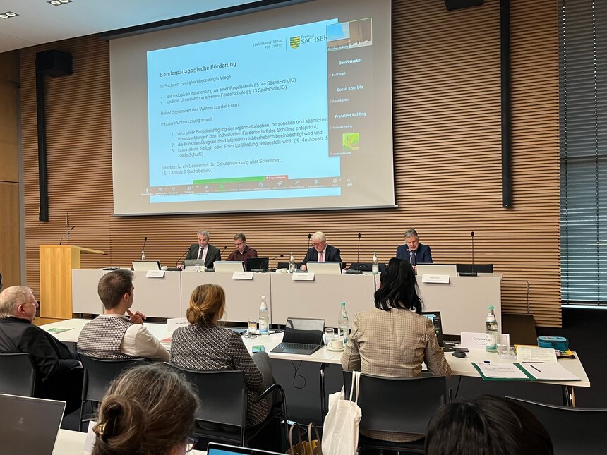 Vier Personen sitzen im Podium, eine Person davon spricht und alle schauen in Richtung Publikum, Betrachter schaut aus Richtung Publikum